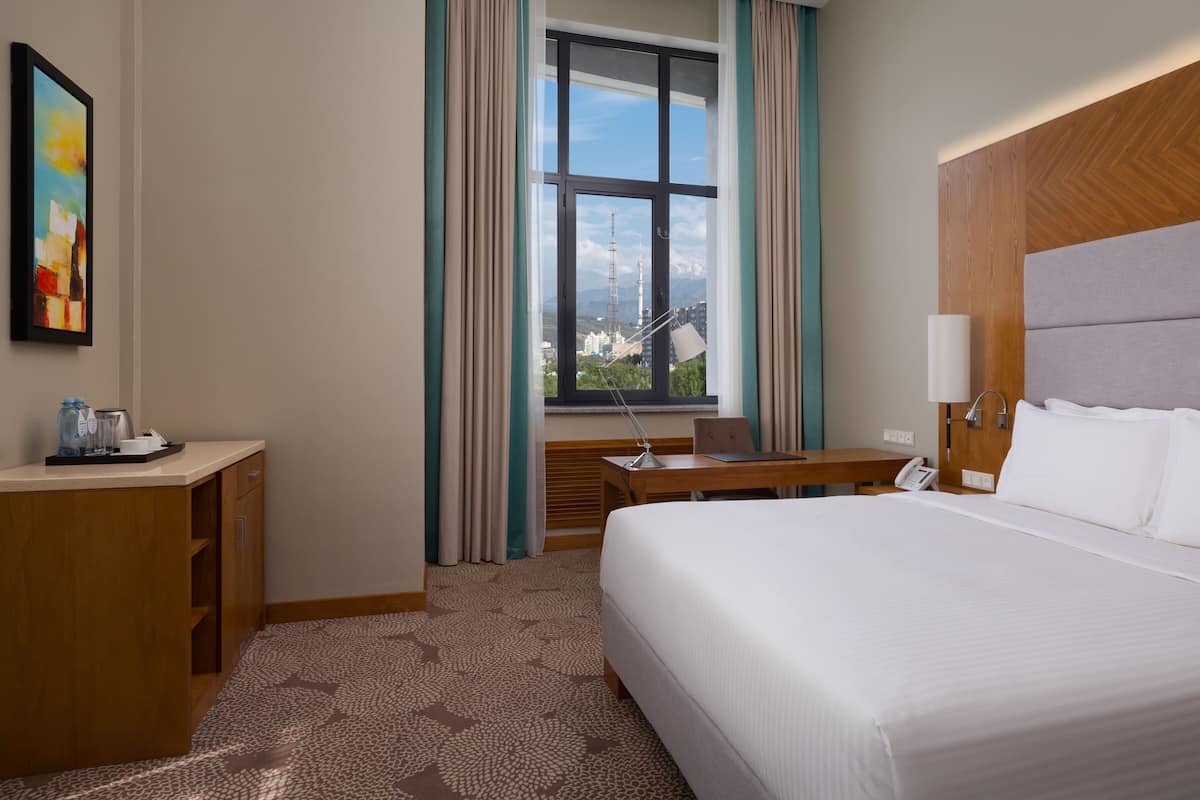 Ett hotellrum med en stor säng, ett skrivbord med en lampa, en telefon och utsikt över stadsbilden genom ett fönster.