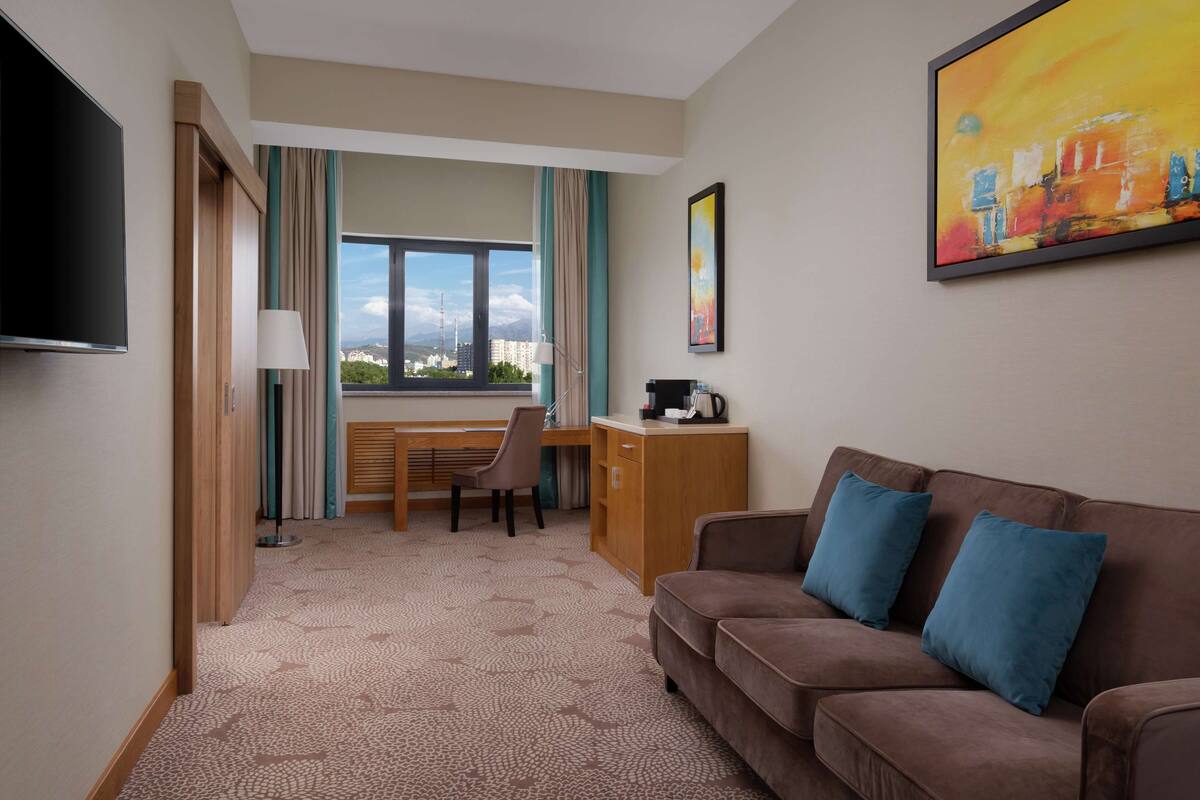 Ett hotellrum med en brun soffa, ett skrivbord, en stol, en tv och en tavla på väggen.