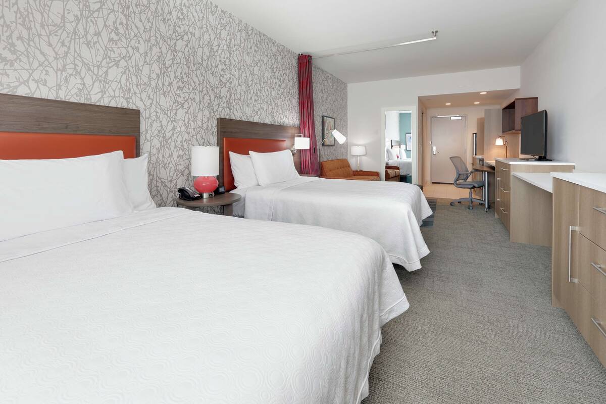 A hotel room with two beds, a desk, a chair, a TV, and a bathroom visible in the background.