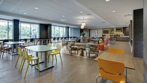 A spacious indoor area with multiple tables and chairs, large windows, and a kitchen area. - Home2 Suites BY Hilton Hagerstown (Hagerstown)