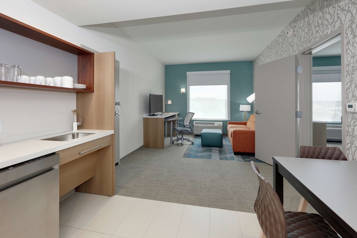 A compact hotel room with a kitchenette, a sofa, a dining table, and a view of the outdoors.