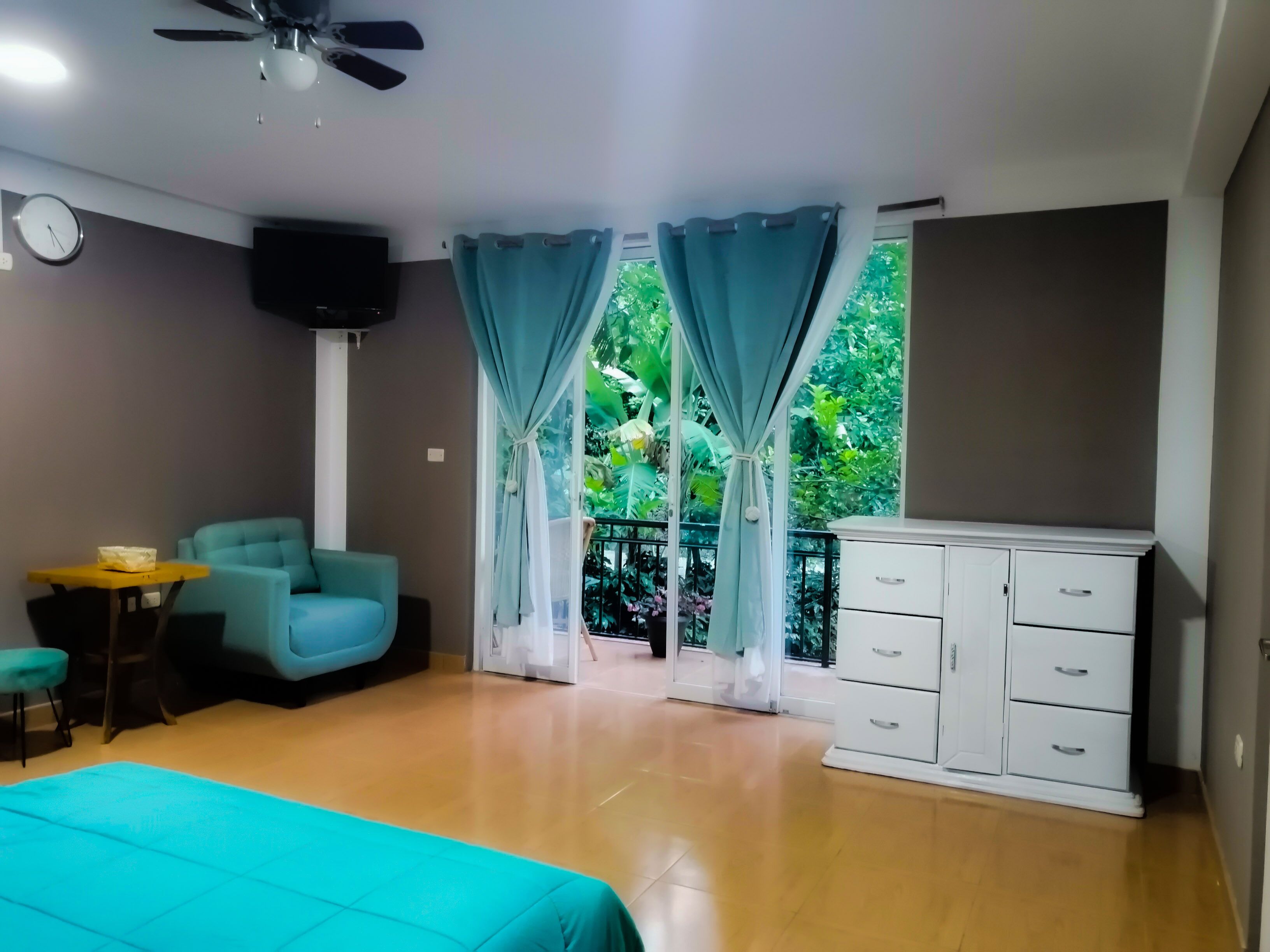 A bedroom with a bed, a desk, a chair, a TV, a wardrobe, and a balcony with curtains.