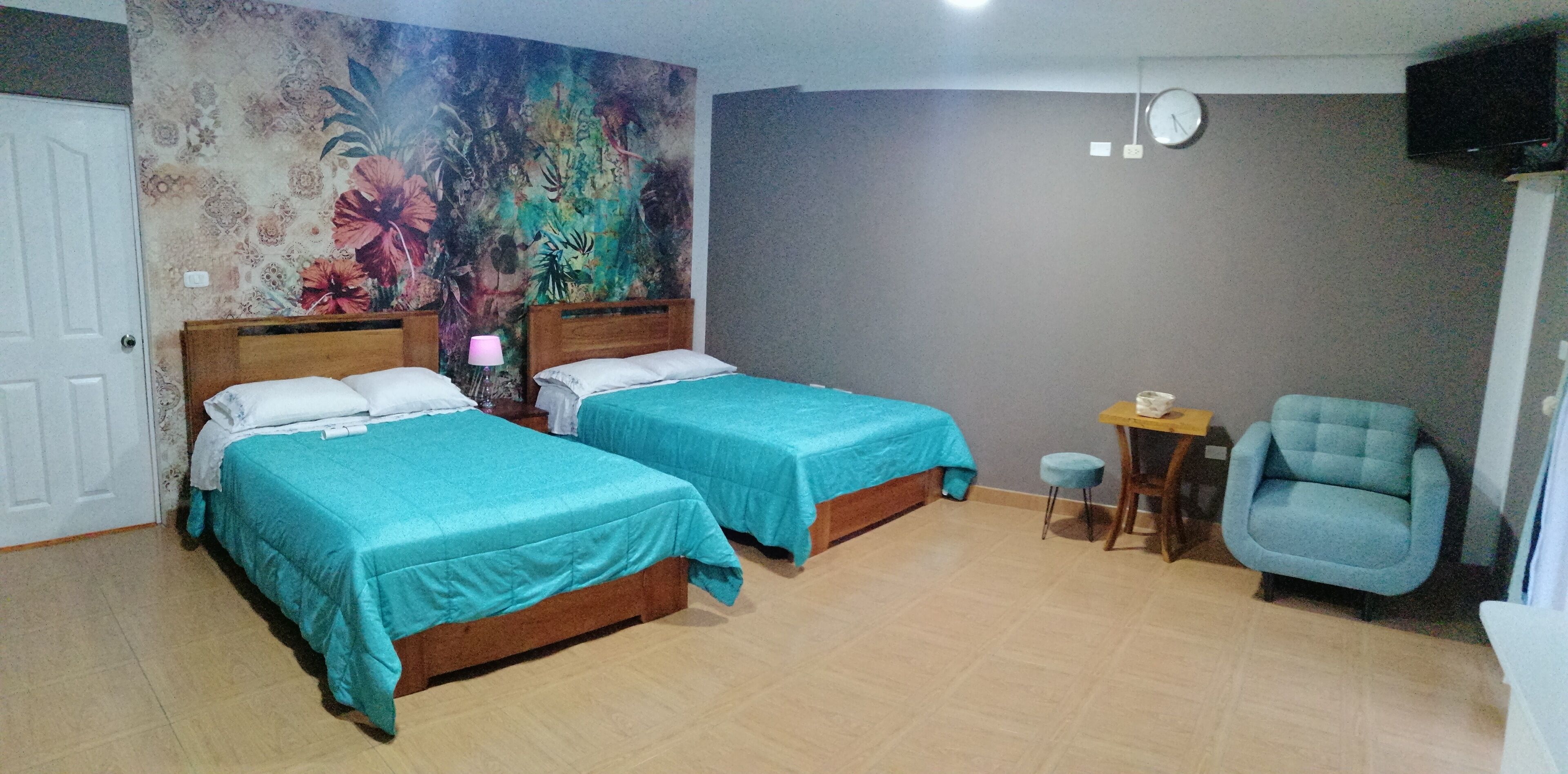A hotel room with two beds, a small table, a chair, a clock, and a floral wall mural.