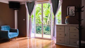 A room with a blue armchair, a television mounted on a white pillar, a cabinet with drawers, and a large window with curtains. - Zoila's Suite Escape (Tingo)