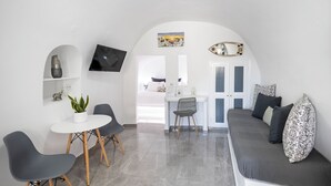 A modern living space with a sofa, dining table, and chairs, a TV mounted on the wall, and a small plant on a shelf. - White Side Suites (Santorini)