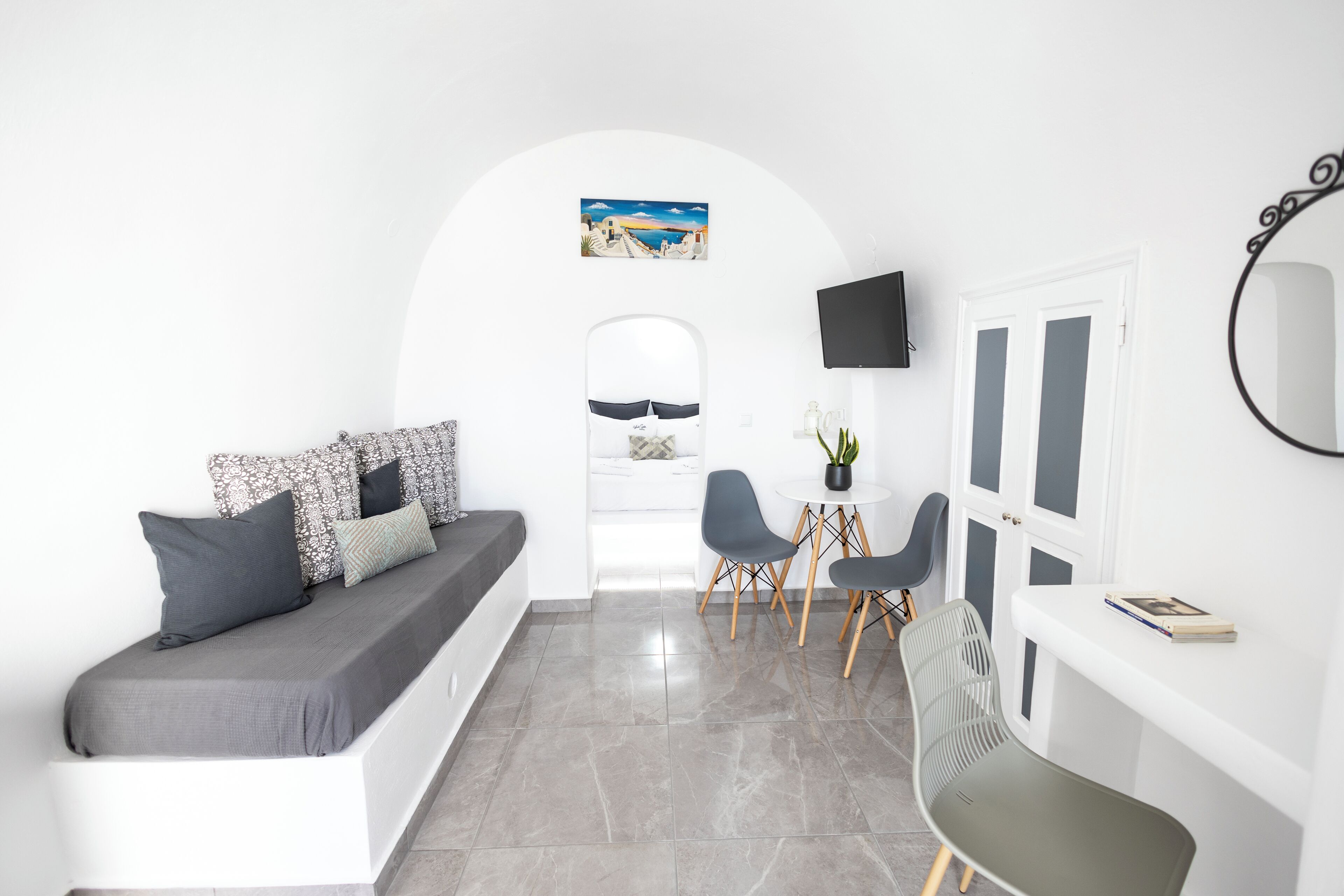 Habitació moderna i minimalista amb un sofà blanc, coixins grisos, taula rodona petita amb dues cadires, televisió de pantalla plana i un mirall gran.