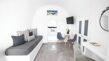 Habitació moderna i minimalista amb un sofà blanc, coixins grisos, taula rodona petita amb dues cadires, televisió de pantalla plana i un mirall gran.