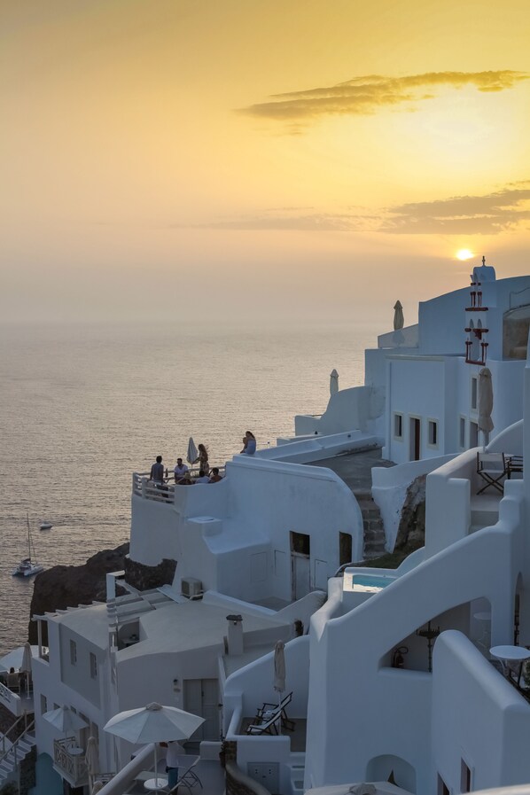 A coastal building with white architecture, a balcony with people, and a view of the sea at sunset. - White Side Suites (Santorini)