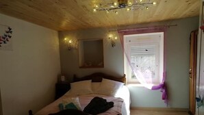 A bedroom with a wooden ceiling, a bed with pillows, a nightstand, a window with curtains, and a mirror on the wall.