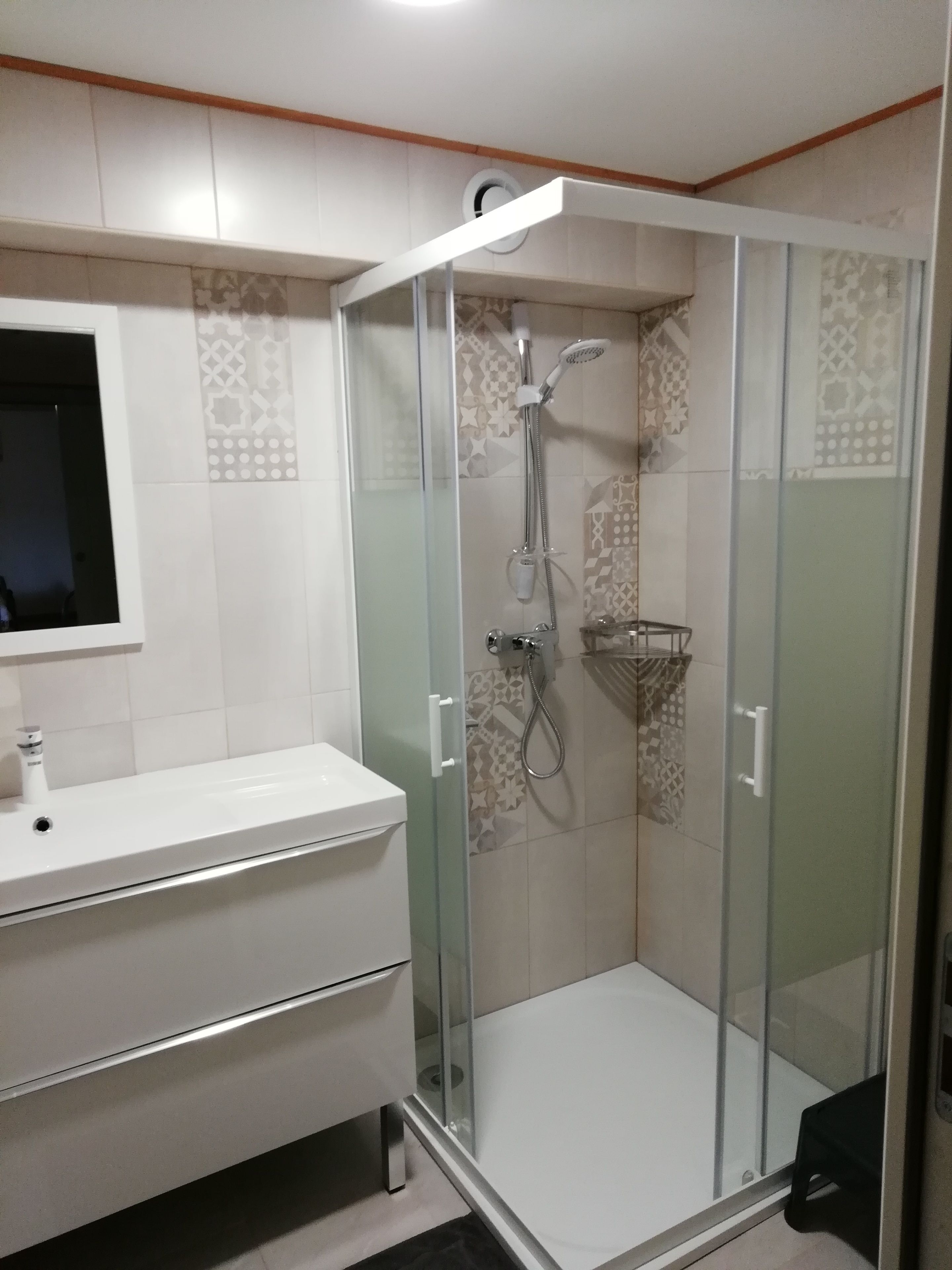 Un baño moderno con ducha envuelta en vidrio, un lavabo blanco y azulejos estampados.