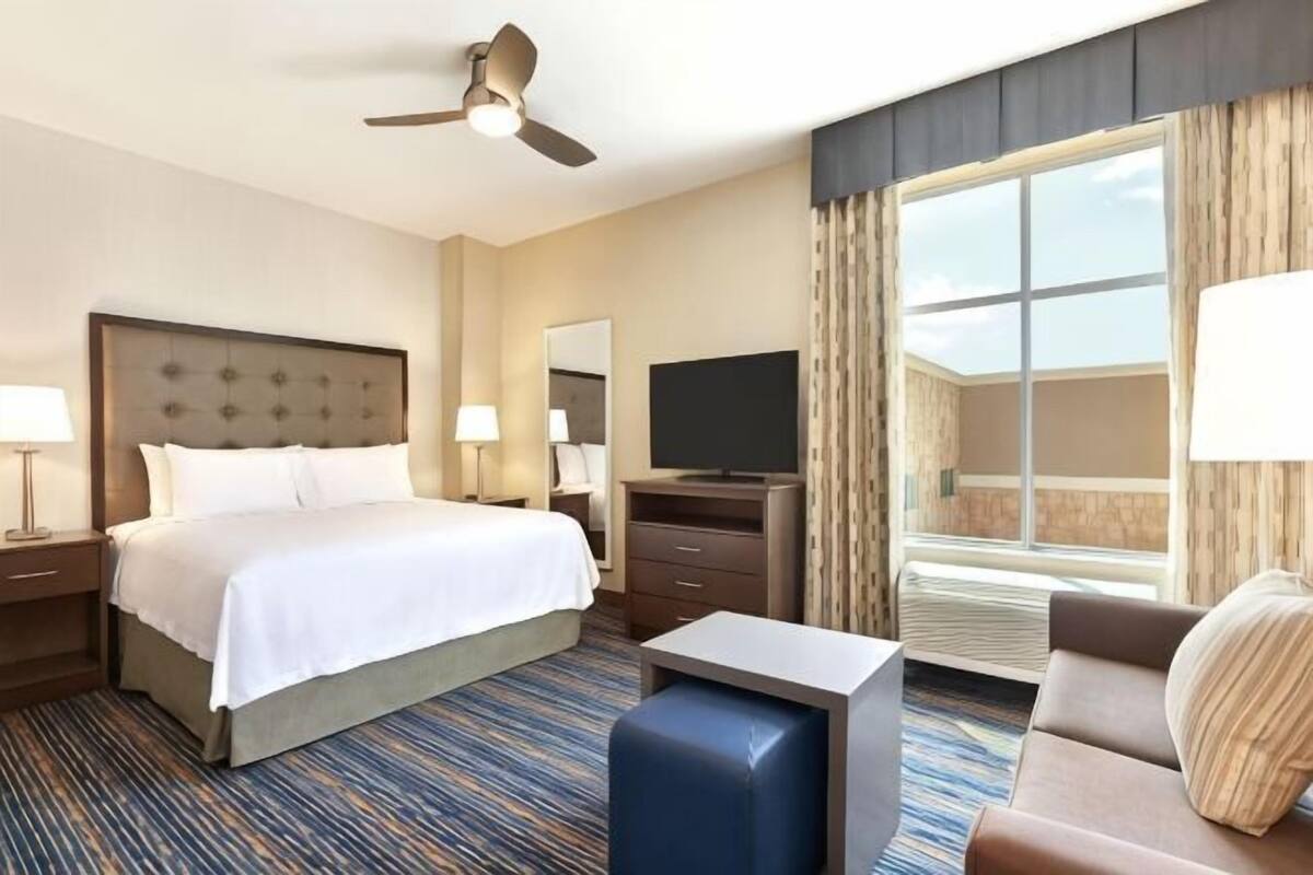 A hotel room with a bed, a desk, a television, a ceiling fan, and a large window.