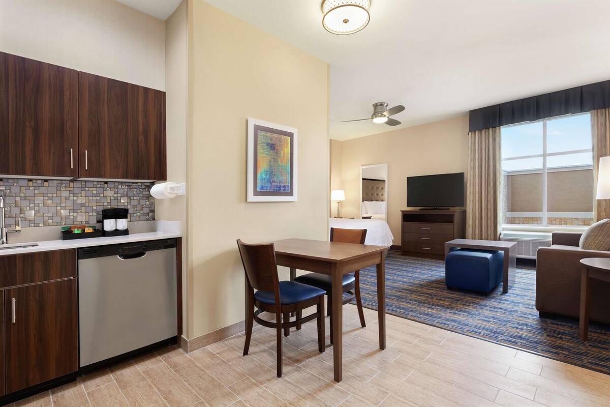 A hotel room with a kitchenette, a dining area with a table and chairs, a living area with a sofa and a TV, and a large window with curtains.