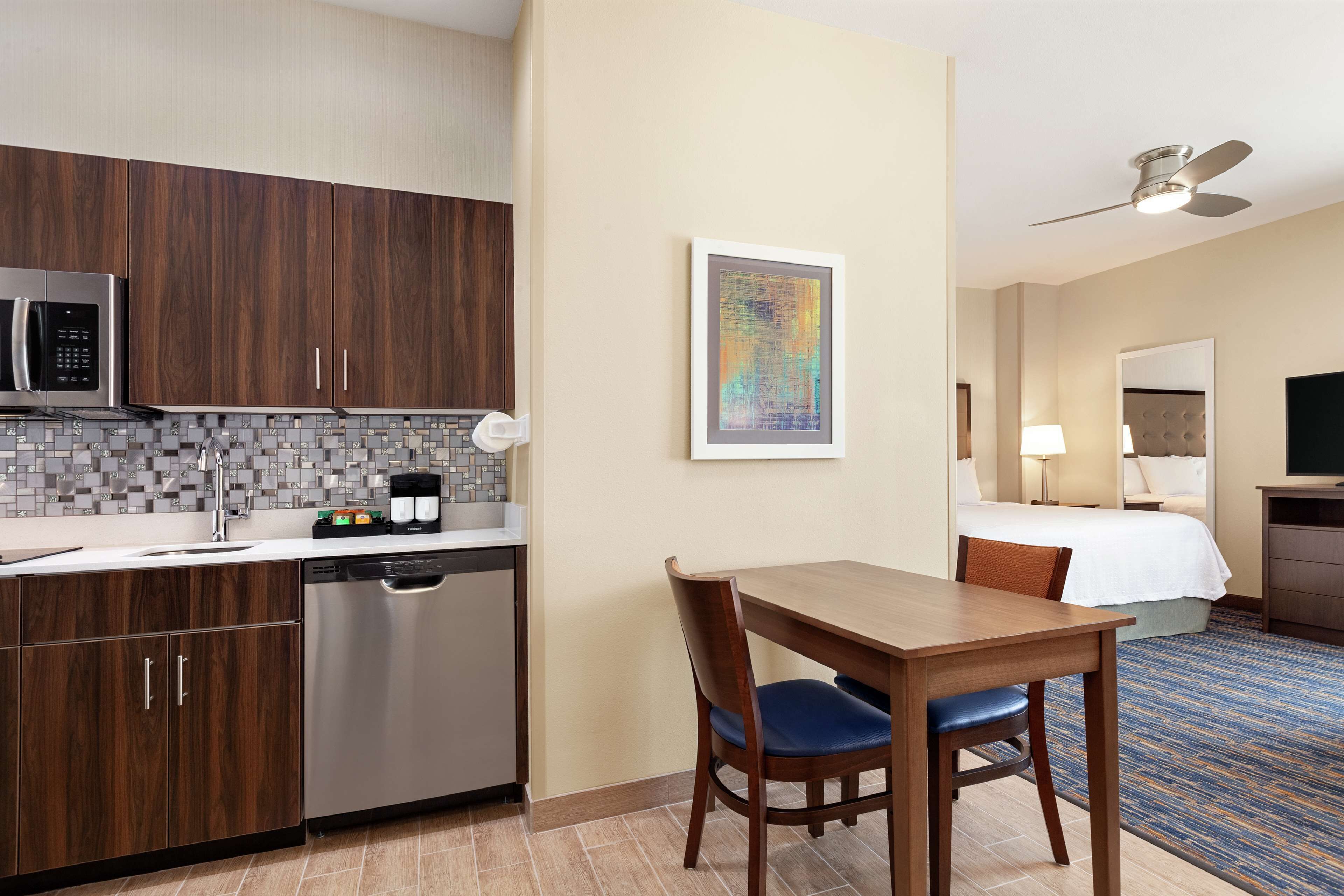 Une chambre d’hôtel équipée d’une kitchenette, d’une table à manger avec des chaises, d’un lit, d’un ventilateur de plafond et d’une œuvre d’art encadrée au mur.
