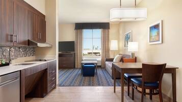 A hotel room with a kitchenette, a dining area with a table and chairs, a sofa, a window with a view, and a lamp.