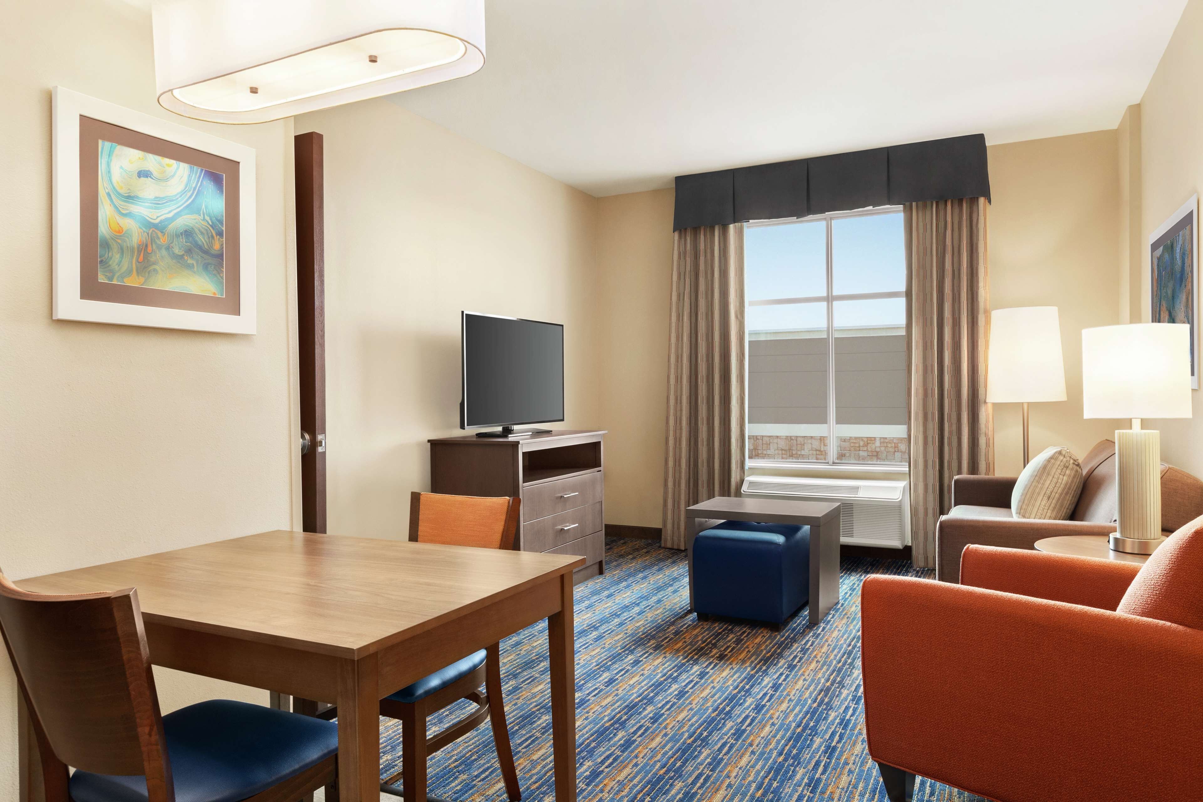Una habitación de hotel con una mesa de comedor de madera, un televisor, un sofá y una ventana con cortinas.