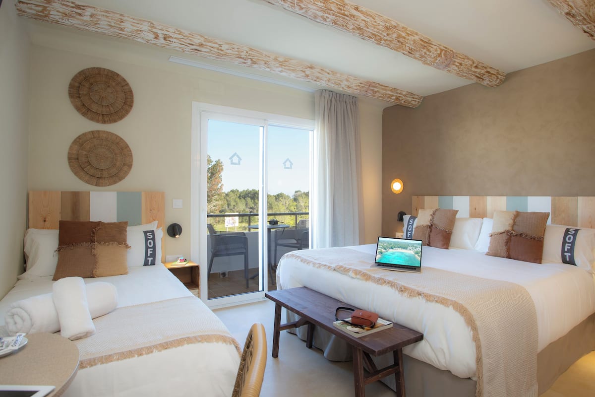 A hotel room with two beds, a laptop on one bed, a table with a laptop and a book, and a balcony with chairs.