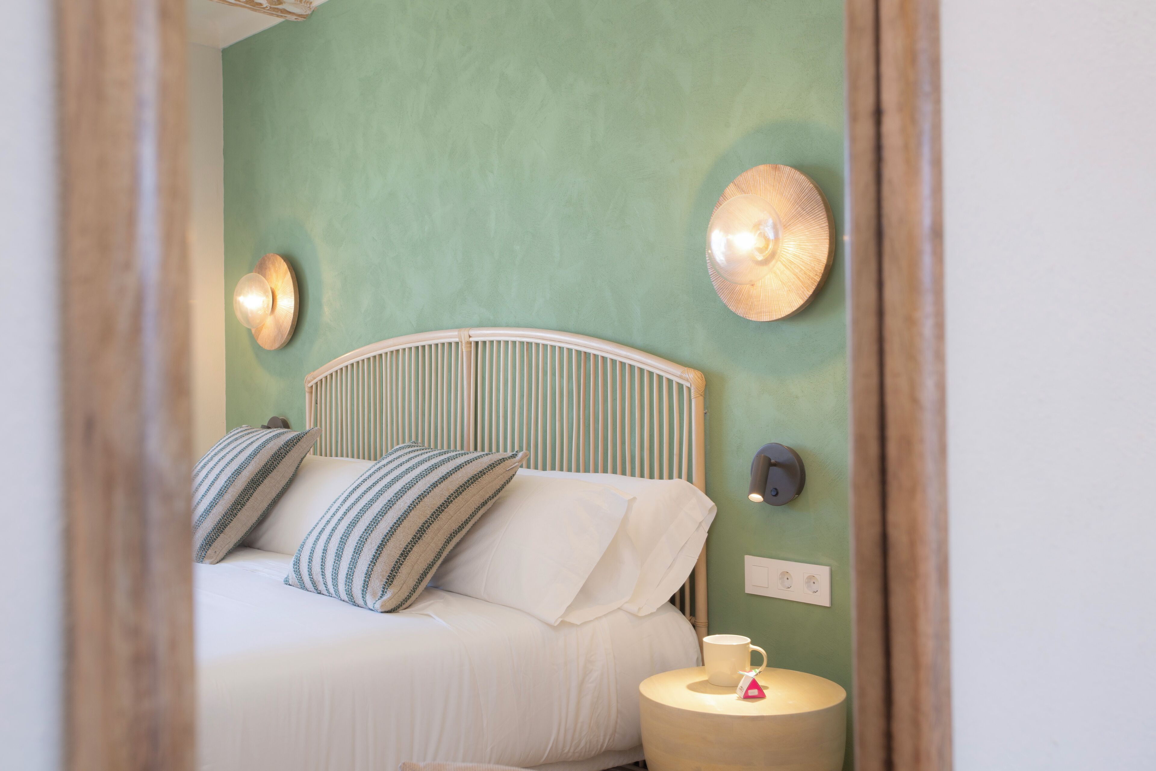 Una cama con ropa de cama blanca y almohadas a rayas, una mesita de noche redonda con una lámpara, y un aplique de pared.