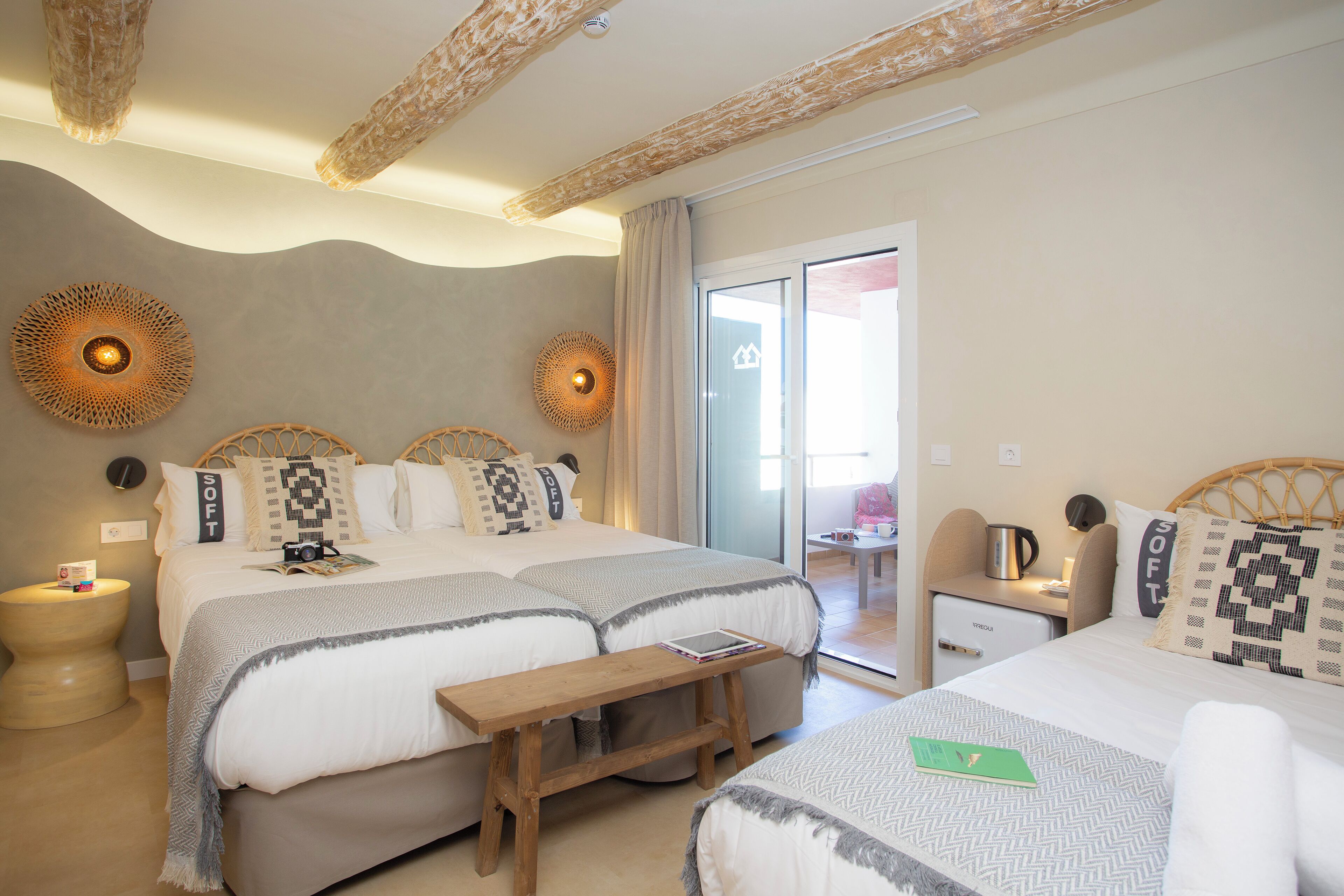 A hotel room with two beds, a wooden bench, a small table with a kettle, and a view of the outside.
