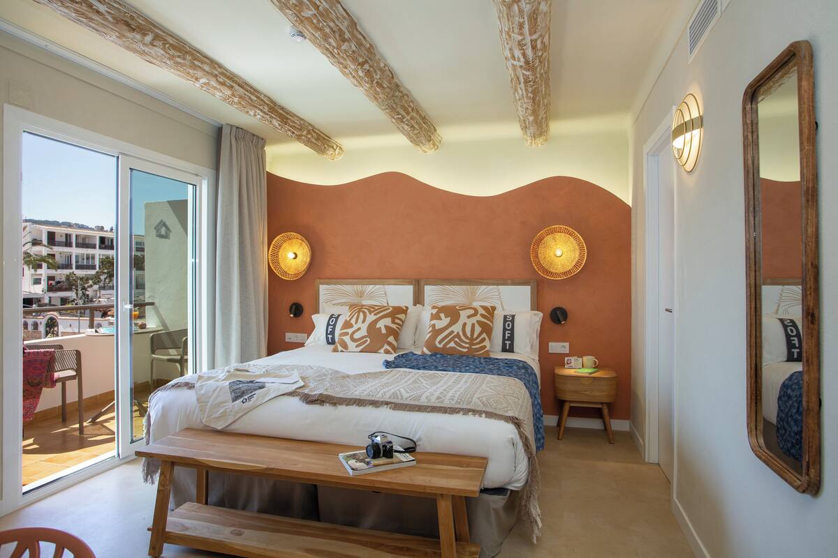 A bedroom with a bed, bedside tables, a bench, a large window, and a view of the outside.