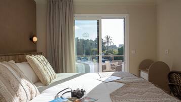 Une chambre d’hôtel avec un grand lit, un appareil photo et un magazine posés sur le lit. Il y a une fenêtre donnant sur des palmiers et une piscine.