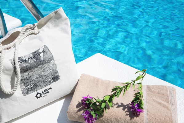 Una bolsa blanca de tela con un diseño e inscripción, una toalla beige doblada y una rama con flores moradas sobre una superficie blanca cerca de una piscina.