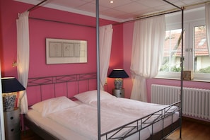 A bed with a metal frame, a bedside table with a lamp, a window with curtains, and a radiator.