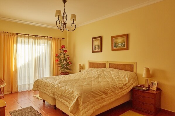 A bedroom with a large bed, a chandelier, two paintings, a potted plant, and a bedside table with a lamp.
