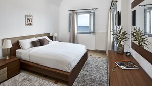 Una camera da letto con un letto, comodino, lampada e vista sul mare attraverso una finestra.