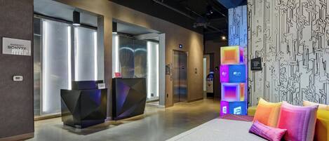 A hotel lobby with a reception desk, elevators, and a seating area with colorful cushions.