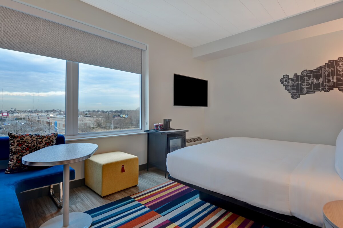 A hotel room with a large bed, a small table, a sofa, a colorful striped rug, a TV on the wall, and a window with a city view.