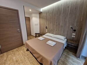 A modern hotel room with a wooden floor, a bed with a beige quilt, two bedside tables, a nightstand, and a wooden accent wall. - Arya Cagliari (Cagliari)