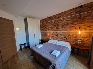 A modern bedroom with a wooden wall, a bed with a brown bedspread, a bedside table, and a hanging light fixture. - Arya Cagliari (Cagliari)