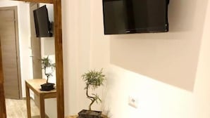 A hallway with a wooden bench, a small potted plant, a television mounted on the wall, and a mirror reflecting the room. - Arya Cagliari (Cagliari)