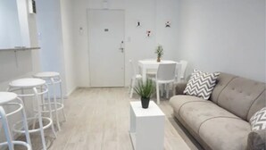 Un salon moderne avec un canapé beige, une table basse blanche agrémentée d’une plante, une table à manger blanche avec des chaises, et un coin bar avec des tabourets blancs.