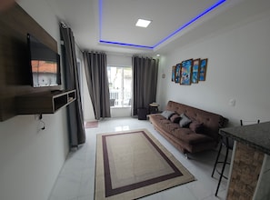 A living room with a brown sofa, a flat-screen TV mounted on a wooden shelf, a patterned rug, and a balcony with curtains. - Apart ZSS (Penha)