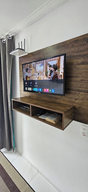 A flat-screen TV mounted on a wooden shelf with a remote control and a power outlet below it. - Apart ZSS (Penha)