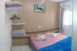 Un dormitorio con cama, almohadas, un estante colgante, un cuadro y un equipo de aire acondicionado.