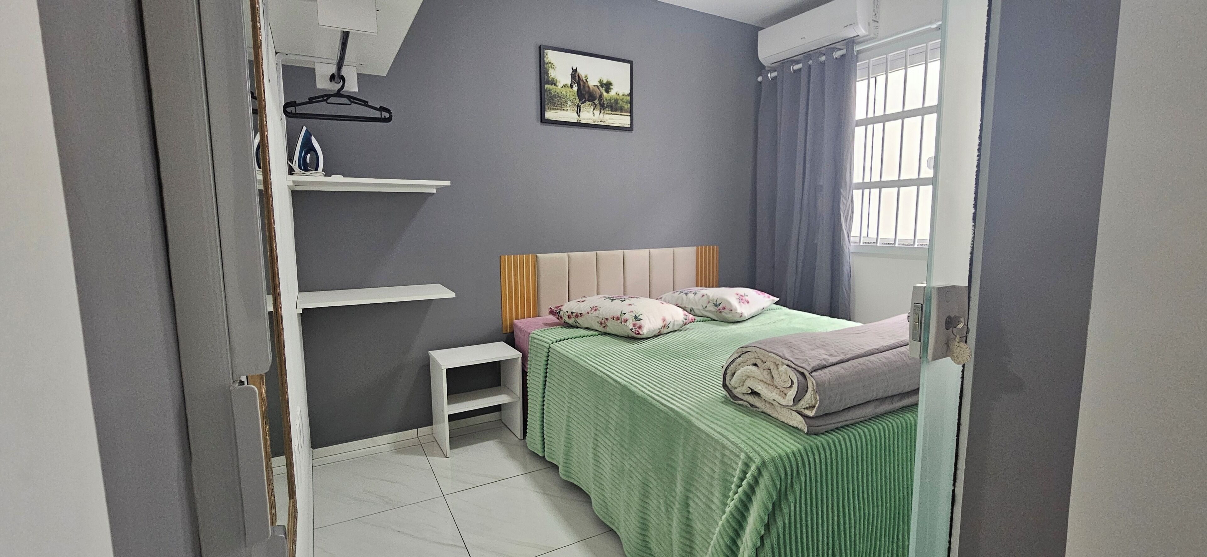 Quarto com cama, almofadas, uma pequena mesa de cabeceira, um ferro de passar roupa e uma janela com cortinas.