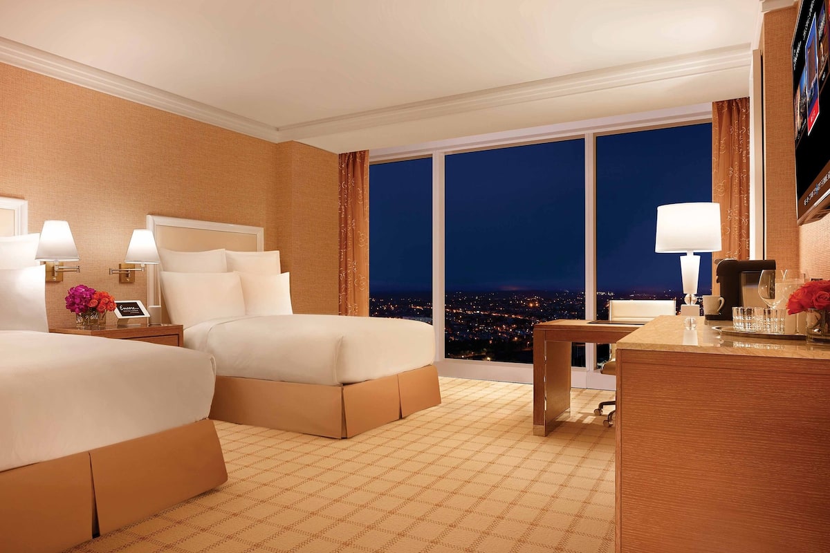 Habitación de hotel con dos camas, un escritorio, una silla, un televisor y un ventanal con vistas a la ciudad.