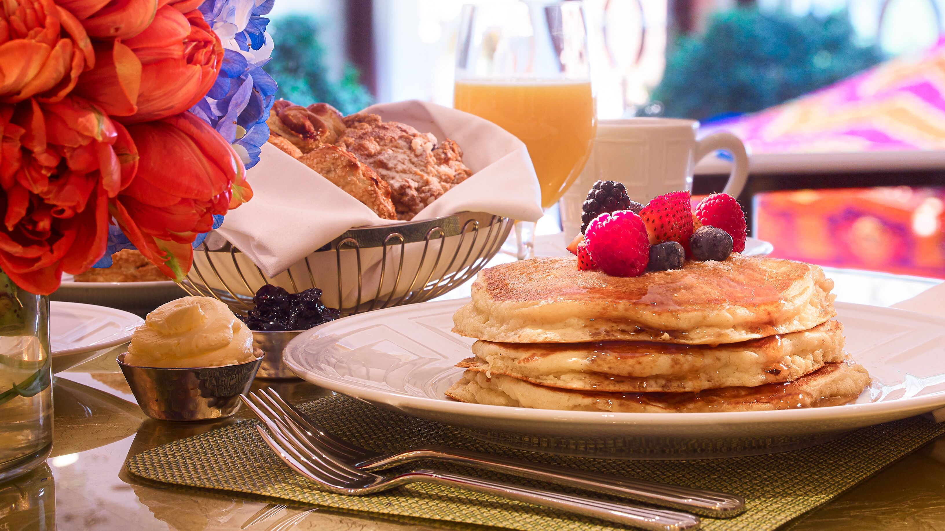Desayuno con tortitas, frutos rojos y zumo de naranja sobre una mesa.