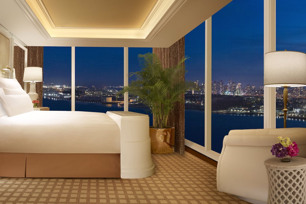 Habitación de hotel con una cama grande, una mesita de noche, una planta en maceta, vista a un paisaje urbano nocturno y una zona de estar con vistas.