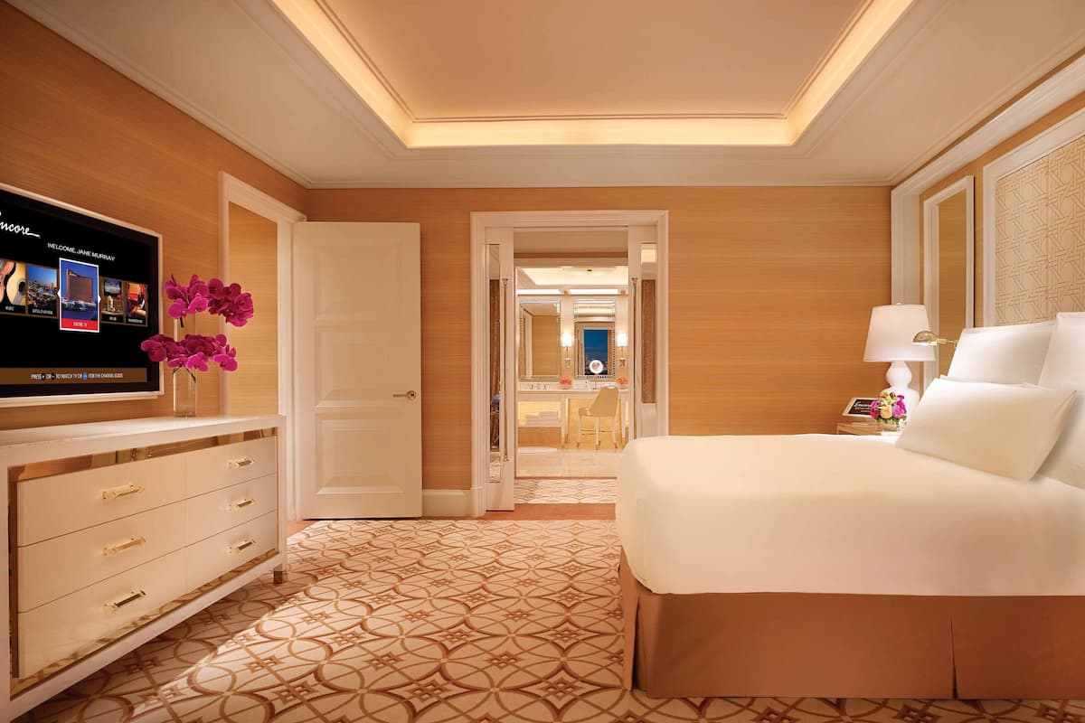Una habitación de hotel con una cama grande, un televisor montado en la pared, un escritorio con una silla y un baño visible a través de una puerta abierta.