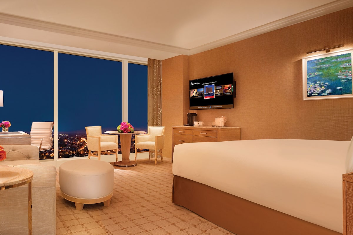 Habitación de hotel con una cama grande, televisión, escritorio con silla, mesita con un jarrón de flores y vistas a la ciudad de noche.