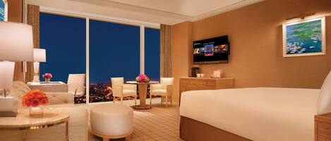 A hotel room with a large bed, a TV, a desk with a chair, a small table with a vase of flowers, and a view of the city at night.