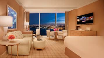 A hotel room with a city view, a TV, a desk, a sofa, a chair, and a bed.