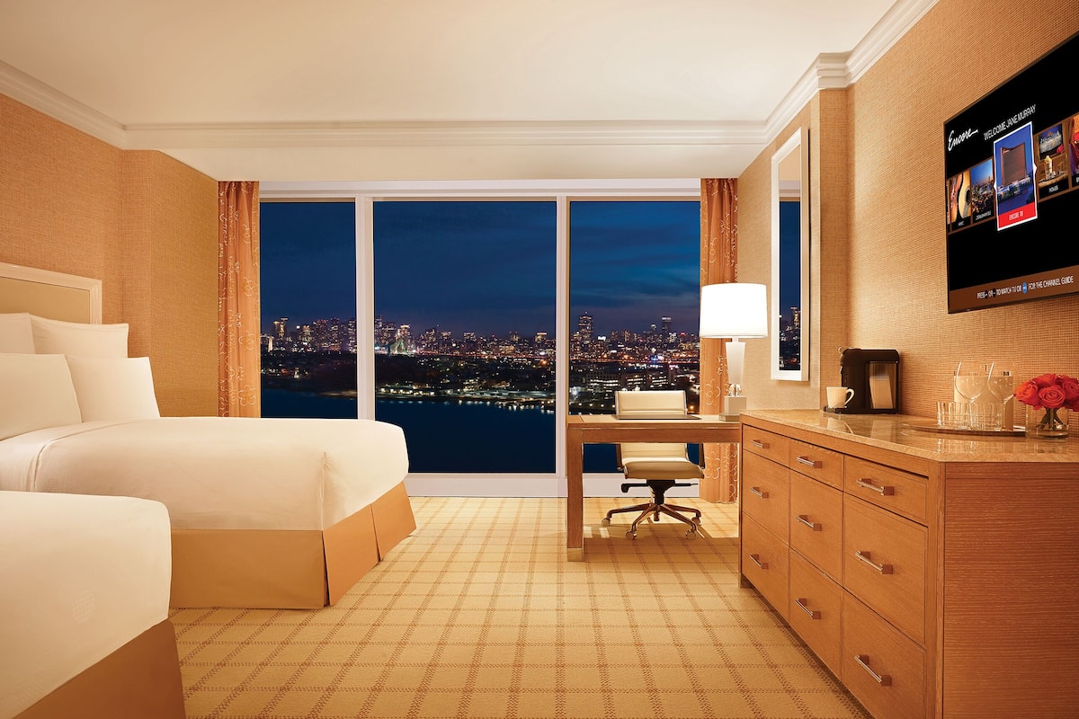 Habitación de hotel con vista a la ciudad, un escritorio, dos camas y un televisor en la pared.
