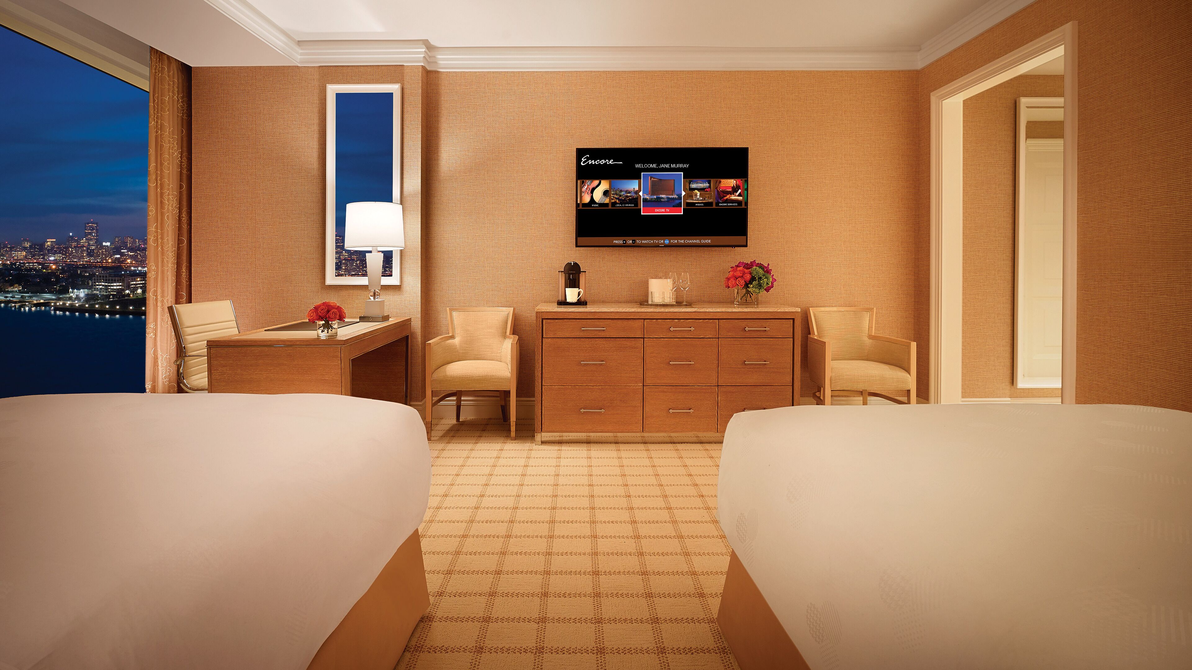 Habitación de hotel con dos camas, un escritorio con lámpara, televisión y vistas a la ciudad.