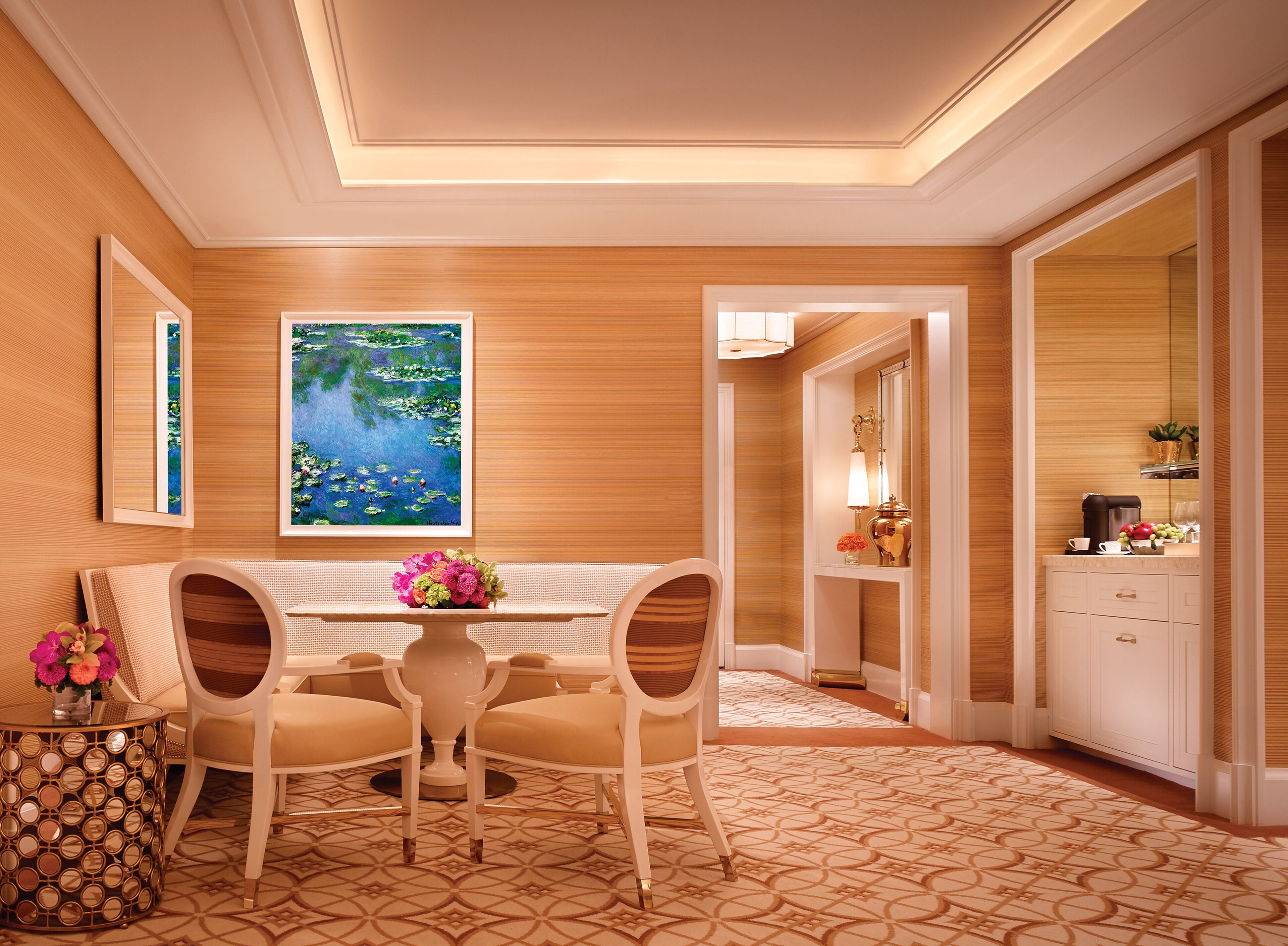 A hotel room with a wooden paneled wall, a large painting of a pond, a dining area with a table and chairs, and a kitchen area with cabinets and a coffee machine.