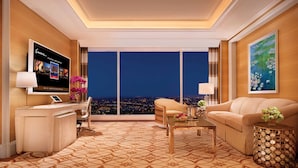 Habitación de hotel moderna con un ventanal grande que ofrece vistas a la ciudad, un televisor de pantalla plana, un sofá, una mesa de centro y un cuadro decorativo.
