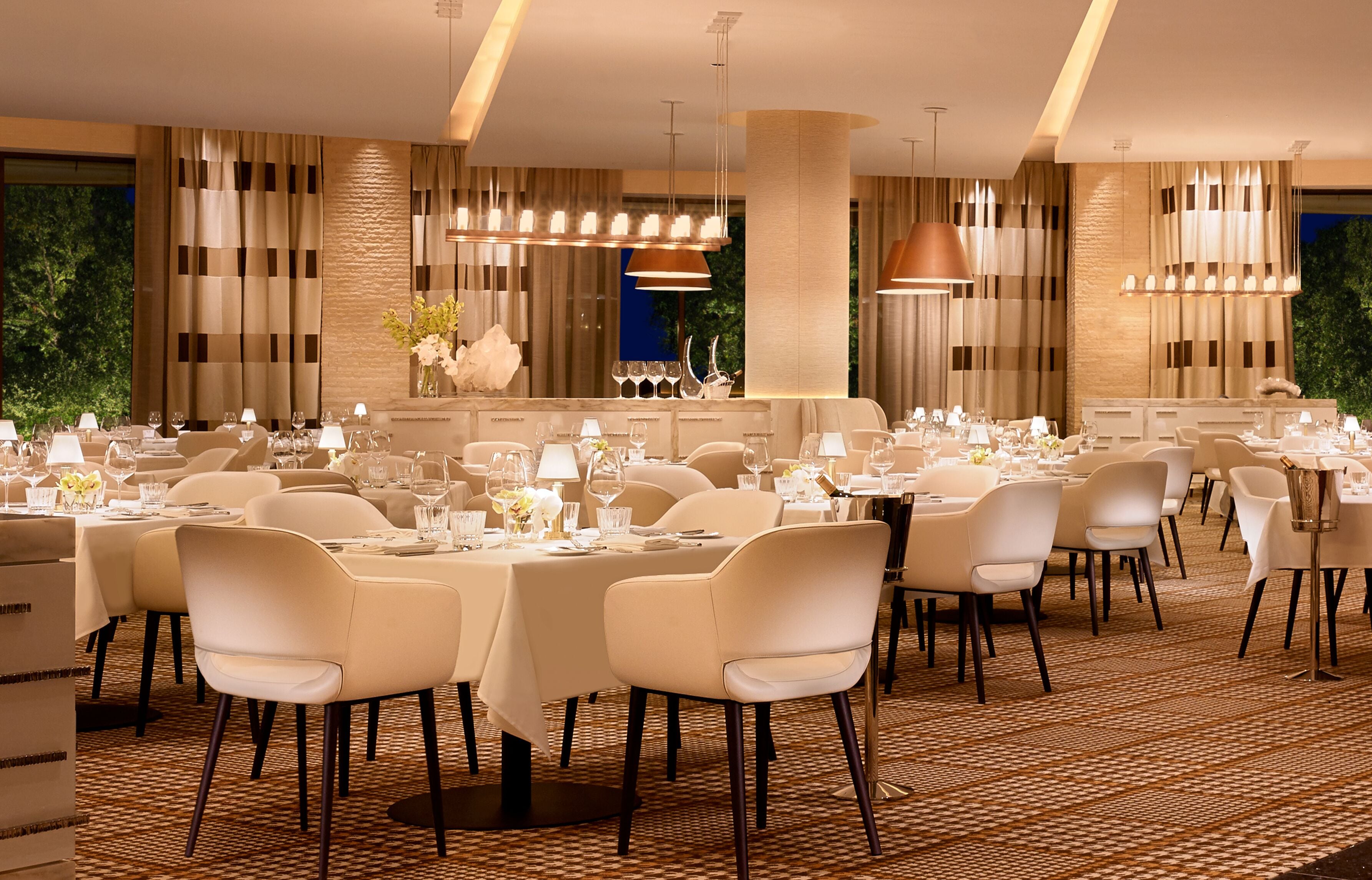 A spacious dining area with multiple tables and chairs, elegant lighting fixtures, and a view of greenery outside.